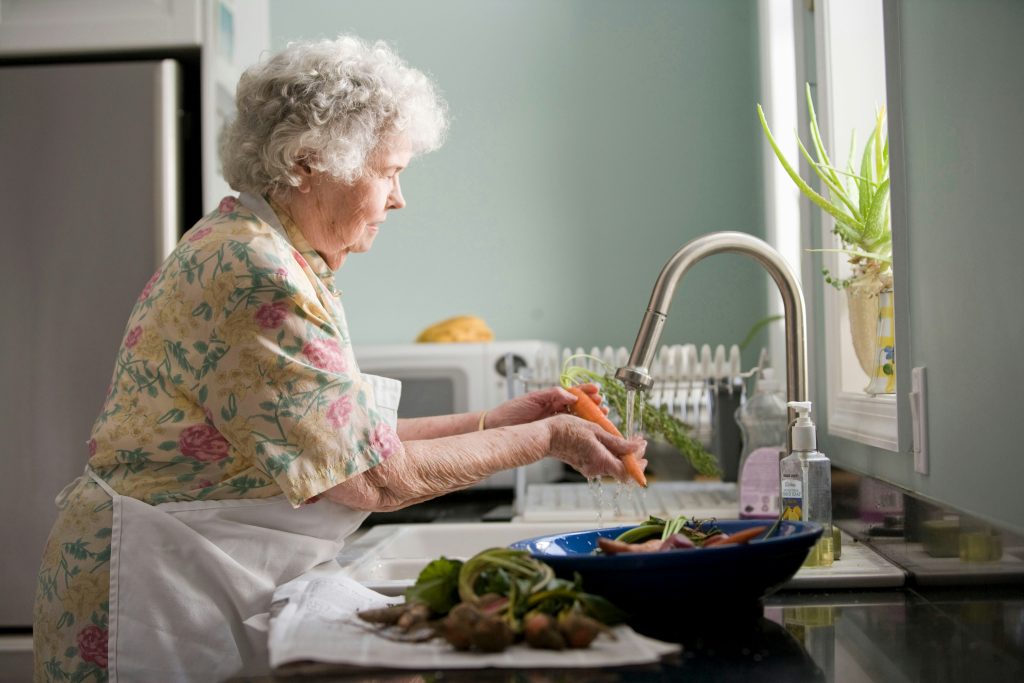 Mujer de la tercera edad vestida con camisa amarilla de flores, lavando verduras, con expresión tranquila, de fondo cocina.
