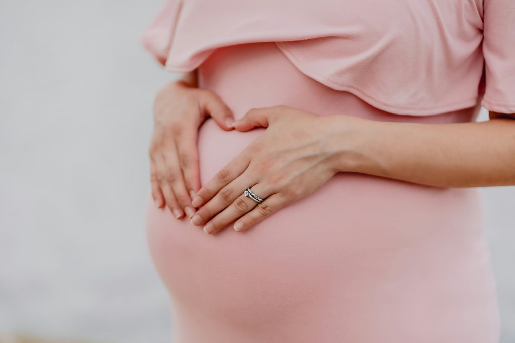 Vientre de mujer embarazada que toma ubiquinol vistiendo un vestido rosa, con las manos apoyadas levemente la panza formando un corazón y luciendo un anillo plateado en uno de sus dedos.